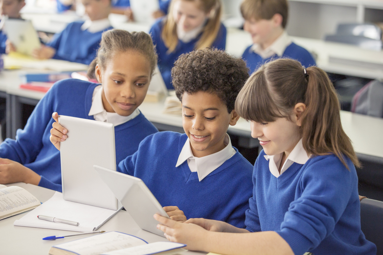 Elementary school children wearing blue school uniforms using digital tablets at desk in classroom